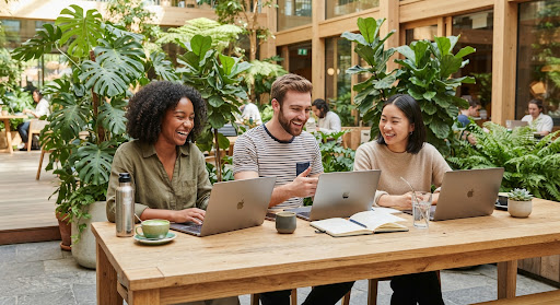 A modern collaborative group of three diverse young professionals working outdoors on laptops at a light oak c